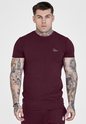 Mężczyzna z tatuażami na szyi i ramionach, ubrany w dopasowaną bordową koszulkę i pasujące spodnie, stojący na jednolitym jasnym tle.