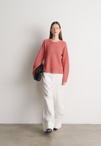 Svetlo roza, rebrasti pleten pulover z širokimi rokavi, v kombinaciji z ohlapnimi belimi hlačami. Oseba drži črno torbico, postavljena v nevtralno ozadje.