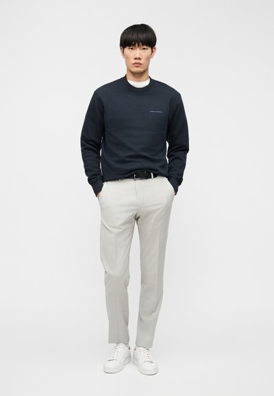 Σκούρο μπλε φούτερ με ριμπ μανσέτες, ανοιχτό γκρι παντελόνια και λευκά αθλητικά παπούτσια. Απλό κεντημένο κείμενο στο στήθος του φούτερ.