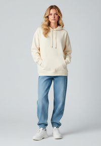 Felpa crema con tasca frontale e cordini, abbinata a jogger in denim azzurro chiaro con polsini elasticizzati e scarpe da ginnastica bianche.