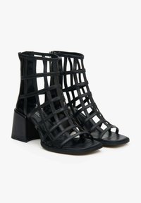 Black heeled sandals with a strappy design and a block heel. Made of smooth material, featuring cut-out details and open toes.