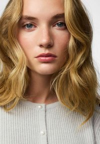 Heledhall hallidega tekstuuriga kampsun, ringkraega ja kahe pärlisonga küljes, seljas inimesel, kelle keskmise pikkusega lainelised blonde juuksed.