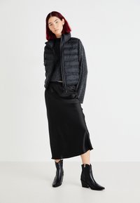 Zwarte puffer vest over een grijze gebreide trui, gecombineerd met een zwarte satijnen rok en zwarte hoge laarzen. Gladde stof, moderne pasvorm en ritsdetails.