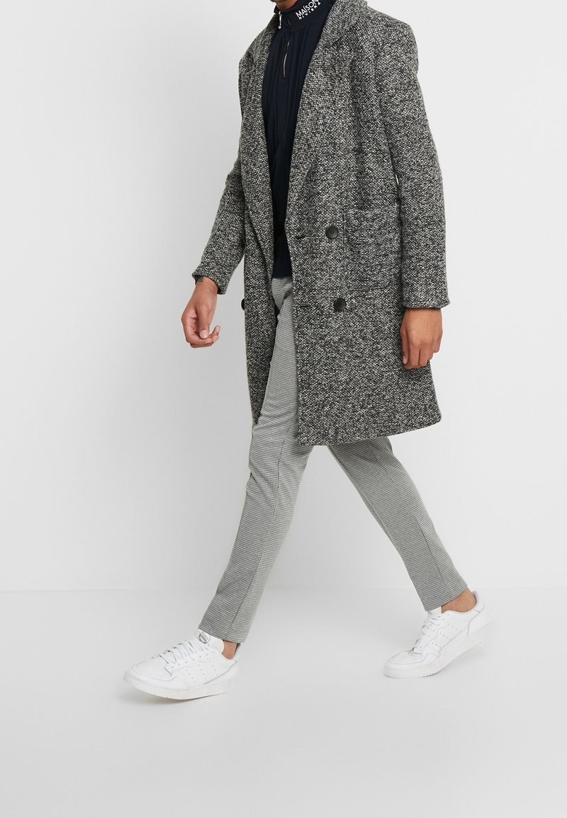 Henkilö kävelee harmaan teksturoidun kaksirivisen takin, mustan vetoketjullisen paidan, harmaiden ruudullisten slim-fit-housujen ja valkoisten lenkkareiden kanssa valkoista taustaa vasten.