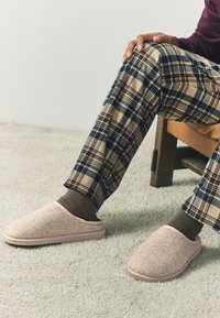 Svetloružové papuče na natiahnutie vyrobené z textúrovaného materiálu so vzorom rybej kosti. Nosené s hnedými ponožkami a károvanými nohavicami na mäkkom koberci.