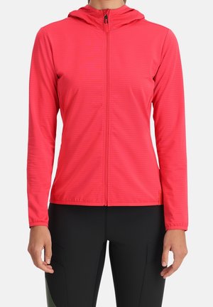 Vrouw draagt een aansluitende, felrode hoodie met rits, lange mouwen en capuchon, gecombineerd met zwarte broek.