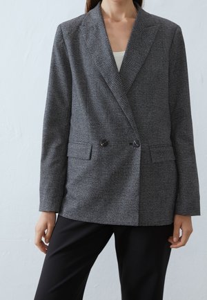 Femme portant un blazer gris à carreaux croisé avec des boutons noirs par-dessus un haut blanc et un pantalon noir sur un fond clair uni.