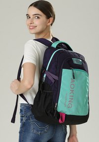 Zaino con una combinazione di materiali viola scuro e teal, compartimenti con zip, spallacci regolabili e un accento rosa nella parte inferiore.