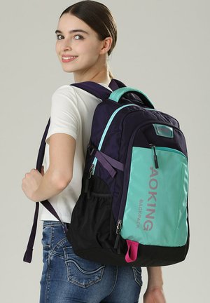 Zaino con una combinazione di materiali viola scuro e teal, compartimenti con zip, spallacci regolabili e un accento rosa nella parte inferiore.