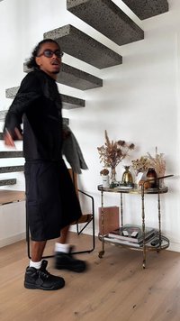 Czarna, oversize koszulka i szorty, czarne sneakersy, teksturowane betonowe schody unoszące się, drewniana podłoga, dekoracyjny stół z suszonymi kwiatami i książkami.