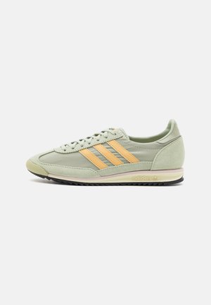 Baskets Adidas vert clair avec des bandes orange, des lacets et une semelle noire texturée, vues du côté extérieur sur fond blanc.