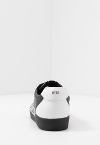 Sneaker bianco e nero con suola nera, tallone bianco con logo metallico "N°21", mostrato da dietro su superficie e sfondo bianchi.