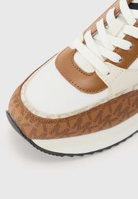 Gros plan sur la pointe d'une basket marron et blanche avec des lacets blancs et un motif marron texturé sur fond blanc.