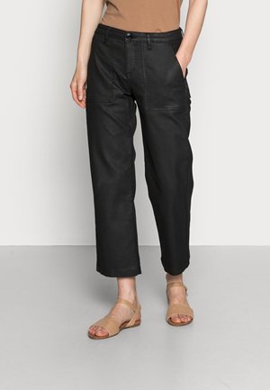 Persoon draagt zwarte cropped broek met grote voorzakken en beige gevlochten sandalen, staand tegen een neutrale achtergrond.