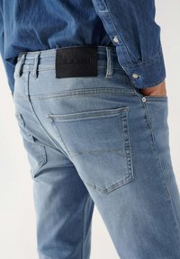 Svijetloplave traperice uskog kroja, s oznakom od crne kože na pojasu, stražnjim džepovima i kontrastnim detaljima šivanja.