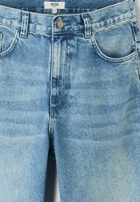 Jean en denim bleu clair avec taille haute, cinq poches, fermeture à bouton métallique et zones légèrement décolorées pour un aspect usé.