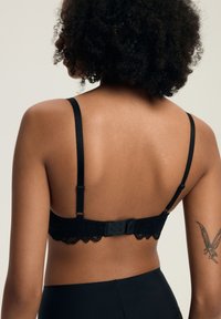 Frau mit lockigem Haar, die einen schwarzen Spitzen-BH und Unterwäsche mit hoher Taille trägt, zeigt ein Elfen-Tattoo auf ihrem oberen linken Arm, von hinten gesehen.