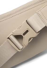 Beige taske lavet af et holdbart syntetisk materiale, med lynlås-lukning og en lys brun rem med en plastik spænde, der sikrer den.