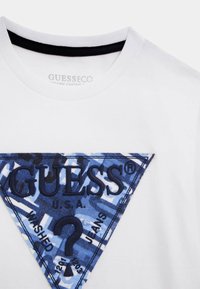 T-shirt bianco in cotone organico con scollo rotondo, caratterizzato da una grafica a triangolo blu con logo "GUESS" ricamato e vari elementi testuali.