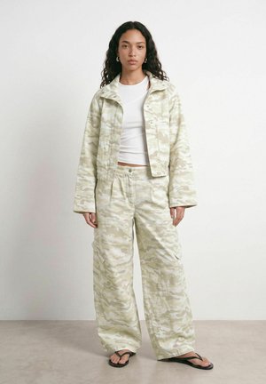 Femme aux cheveux bouclés portant une veste et un pantalon camouflage beige clair avec un haut blanc et des tongs noires, debout devant un fond blanc.