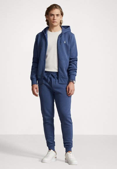 Μπλε βαμβακερή φούτερ με κουκούλα και μπροστινές τσέπες, συνδυασμένο με αντίστοιχες φόρμες. Λευκά αθλητικά παπούτσια ολοκληρώνουν την εμφάνιση. Απλός, καθημερινός σχεδιασμός.