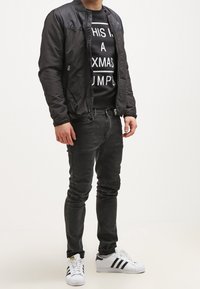 Czarna kurtka bombka z zamkiem, nałożona na czarną bluzę z białym napisem. Slim fit czarne dżinsy i białe trampki z czarnymi paskami dopełniają stylizację.