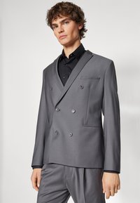 Dobbeltradet grå blazer med en glatt tekstur, har svarte knapper og en kontrasterende svart krage, paret med matchende bukser.