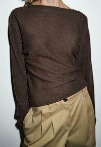 Personne portant un pull à manches longues marron foncé rentré dans un pantalon beige plissé taille haute, les mains dans les poches.