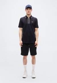 Μαύρο πόλο μπλουζάκι με λευκή αντίθεση στις λεπτομέρειες. Συνδυασμένο με μαύρο σορτς και λευκά αθλητικά παπούτσια. Το μοντέλο φοράει μπλε καπέλο και ανοιχτόχρωμες κάλτσες.