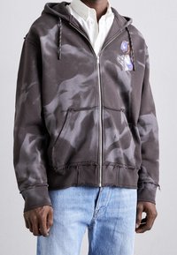 Sudadera con capucha y cremallera en gris oscuro con un patrón desvanecido, bordes sin acabar, bolsillos frontales y un gráfico floral en el lado izquierdo del pecho.