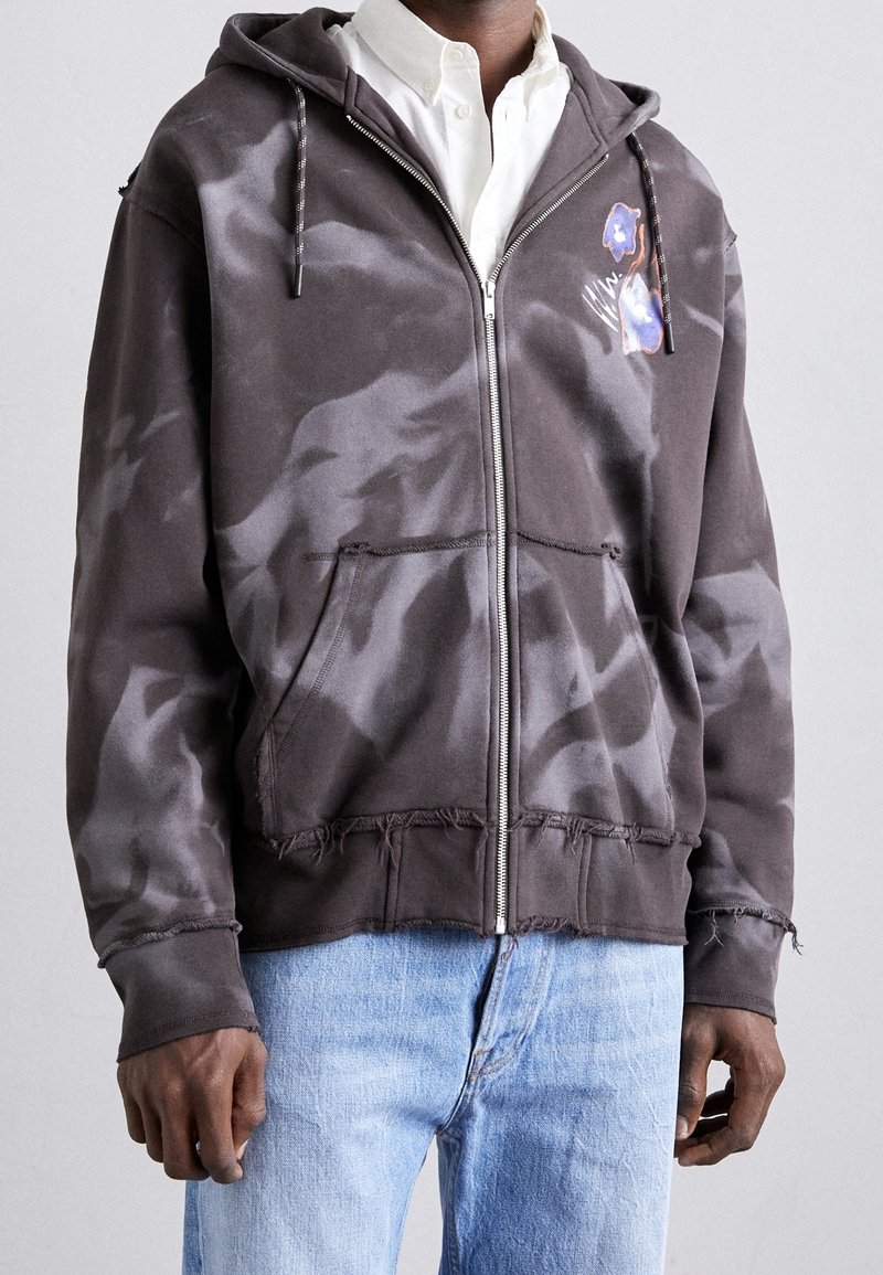 Sudadera con capucha y cremallera en gris oscuro con un patrón desvanecido, bordes sin acabar, bolsillos frontales y un gráfico floral en el lado izquierdo del pecho.