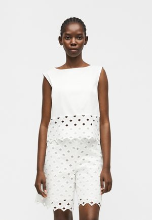 Γυναίκα που φοράει άσπρο αμάνικο τοπ και αντίστοιχα άσπρα σορτς με γεωμετρικά ανοίγματα, στέκεται μπροστά σε ανοιχτό γκρι φόντο.