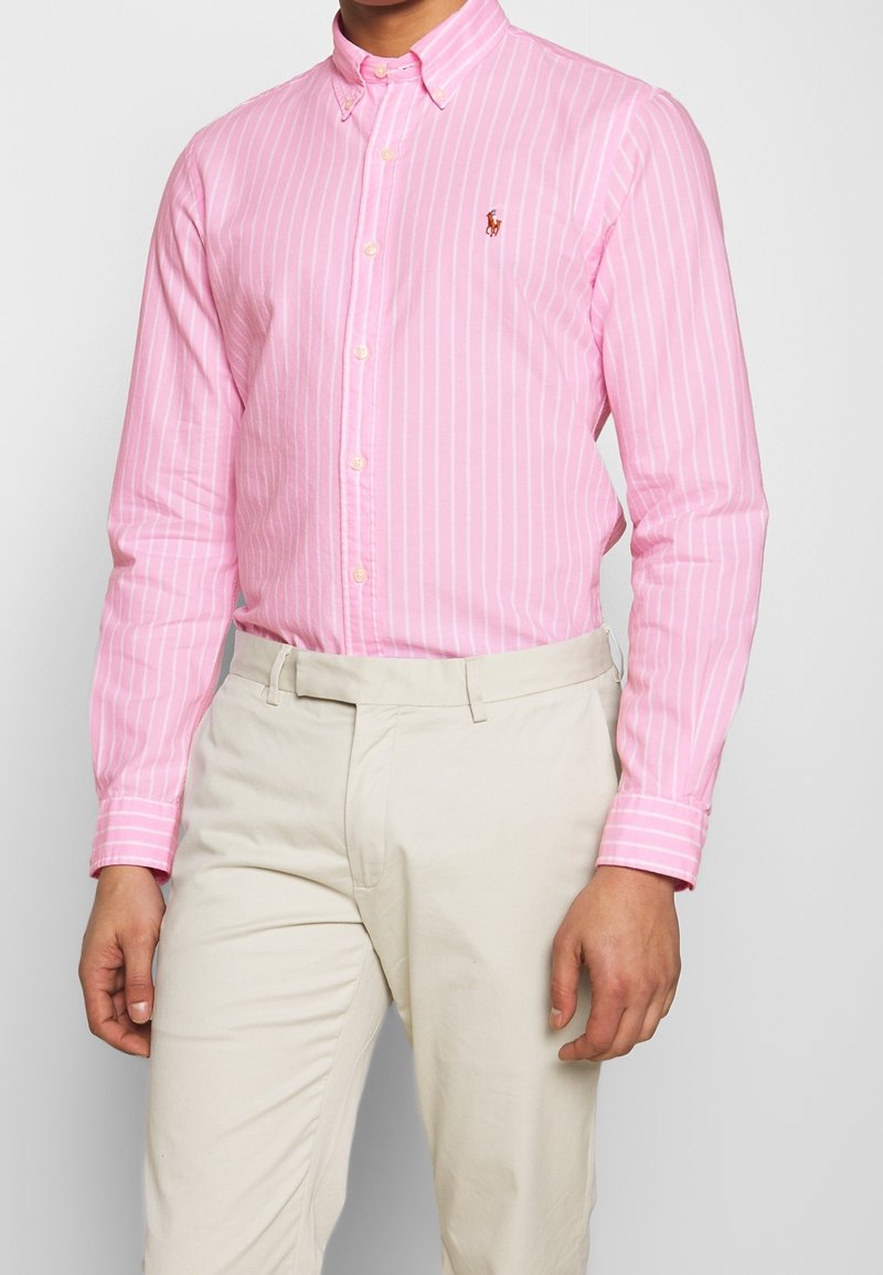 Ljusrosa skjorta med knappar, försedd med vertikala vita ränder, en klassisk krage och en liten broderad logotyp på bröstet. Matchad med beige byxor.