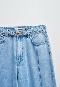 Vaqueros de mezclilla azul claro con un diseño de pierna ancha, bolsillos frontales y un cinturón decorativo con herrajes dorados y una etiqueta que dice "Salsa Jeans".
