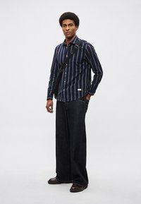 Granatowa koszula w paski zapinana na guziki z białymi i pomarańczowymi liniami, zestawiona z luźnymi, ciemnymi jeansami i brązowymi butami. Do tego czarna torebka na ramię.