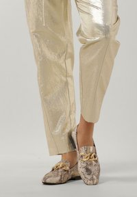 Slangenprint loafers met een gouden kettingaccent, gecombineerd met glanzende, lichtgouden broek. De schoenen hebben een ronde neus en een platte zool.