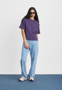 Violeta kokvilnas T-kreklu ar īsām piedurknēm un apļa kakla izgriezumu, saskaņojot ar gaiši ziliem vertikāli svītrainiem biksēm. Modelis valkā tumšus saulesbrilles.