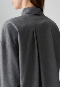 Gray collared shirt with a textured fabric, featuring a back pleat and structured shoulders, emphasizing a relaxed fit.