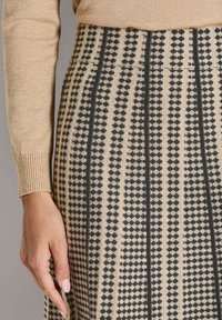 Beige und schwarz senkrecht gestreifter Rock mit geometrischem Muster, getragen mit einem beigen, gerippten Pullover mit Bündchen, entspannt an der Seite hängen.