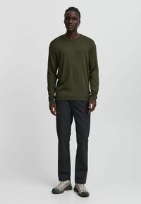 Olijfgroen gebreide trui met lange mouwen, ronde halslijn, geribde zoom, gecombineerd met donkergrijze broek en lichtgrijze sportschoenen.