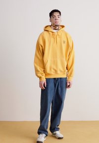 Un sweat à capuche jaune avec une poche avant et un cordon de serrage, associé à un jean bleu ample. Des baskets blanches avec des accents noirs, se tenant devant un fond neutre.