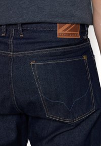 Tmavě modré džíny z denimu s texturovaným povrchem, s oranžovým šitím a hnědou koženou nášivkou s nápisem "Pepe Jeans" na zadní části pasu.