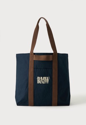 Sac cabas bleu marine avec poignées et bandoulières marron, poche avant, et logo blanc "PMW" avec un graphisme de feuille.