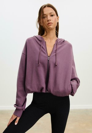 Purple zip-up hoodie with a hood, relaxed fit, dropped shoulders, and ribbed cuffs. Worn over black leggings, featuring a textured fabric.