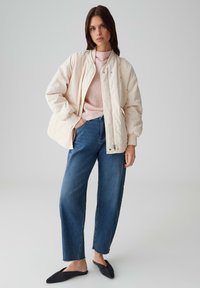 Woman wearing a light beige quilted jacket, pink knit sweater, blue wide-leg jeans, black pointed flats, and carrying a beige shoulder bag.