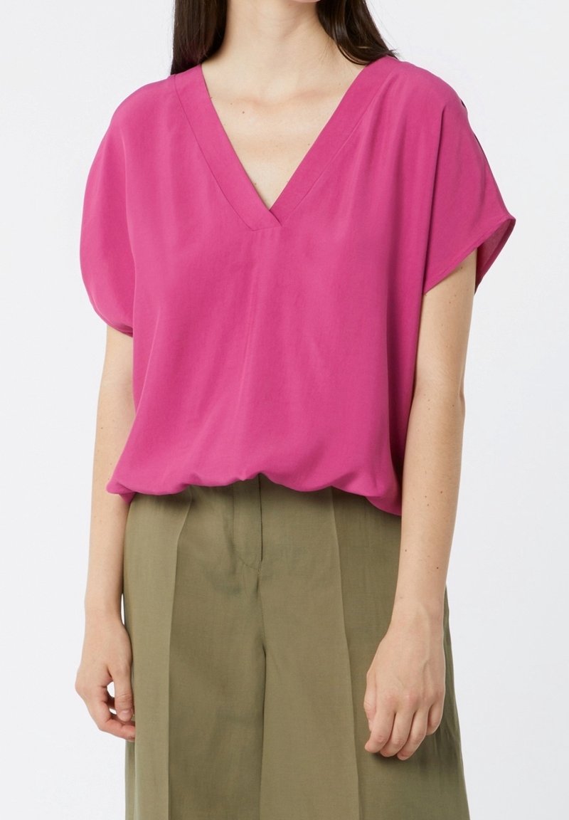 Femme portant un chemisier ample fuchsia à manches courtes et col en V, rentré dans un pantalon taille haute vert olive.