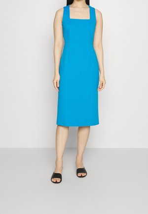 Vrouw draagt een mouwloze, knielange felblauwe jurk met een vierkante halslijn en zwarte open teen slippers, staand tegen een effen witte achtergrond.