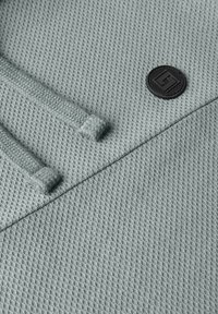 Detailní záběr na texturovanou šedou látku se dvěma šňůrkami a malou černou kulatou nášivkou.