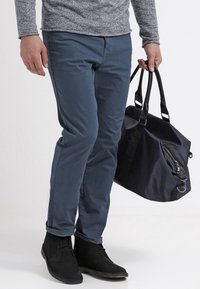 Blauwe katoenen broek met een slim fit, gecombineerd met zwarte suède enkellaarzen. Een marineblauwe duffelbag met een rits en hengsels wordt in één hand vastgehouden.