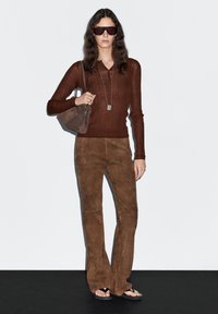 Hnědý žebrovaný top s dlouhým rukávem ve spojení s hnědými semišovými rozšířenými kalhotami a černými žabkami. Modelka je doplněna slunečními brýlemi a malou kabelkou.